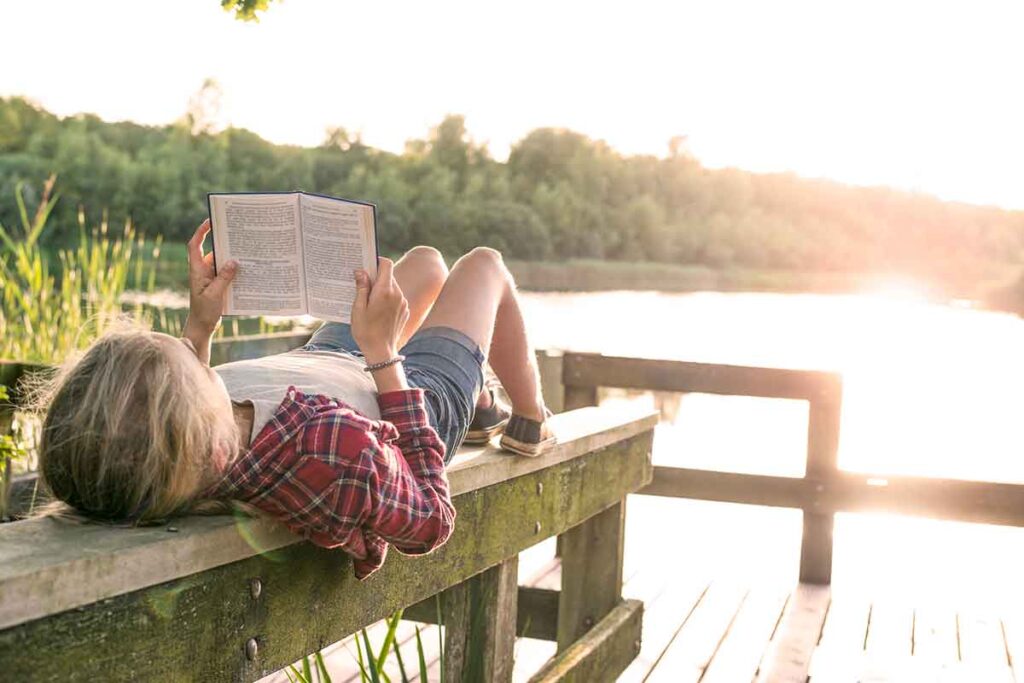 Support Your Teen's Mental Health During Summer Break, teenage girl laying on a dock reading surrounded by water and trees
